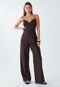 Žena stojící sebevědomě, oblečená v tmavě hnědém bezramínkovém overal s tenkými ramínky a širokými nohavicemi, černých sandálech a masivních náramcích.