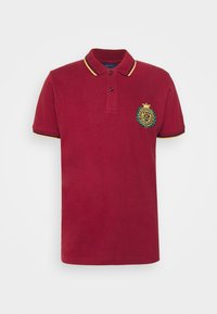 Červené polo tričko z bavlny, s límečkem se žlutými a černými pruhy, dvěma knoflíky a vyšitým emblémem na hrudi.