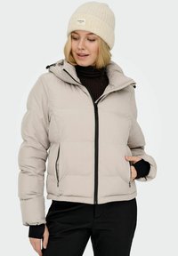Beżowa kurtka puchowa z kapturem, zapinana na zamek, z czarnymi prążkowanymi mankietami i kieszeniami z przodu. Nosi ją osoba z jasnym, dzianinowym beanie.
