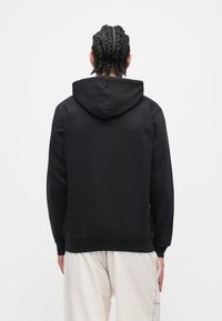 Asmuo su pynėmis, dėvintis juodą džemperį su kapišonu ir šviesių spalvų kelnes, atsisukęs į baltą foną.