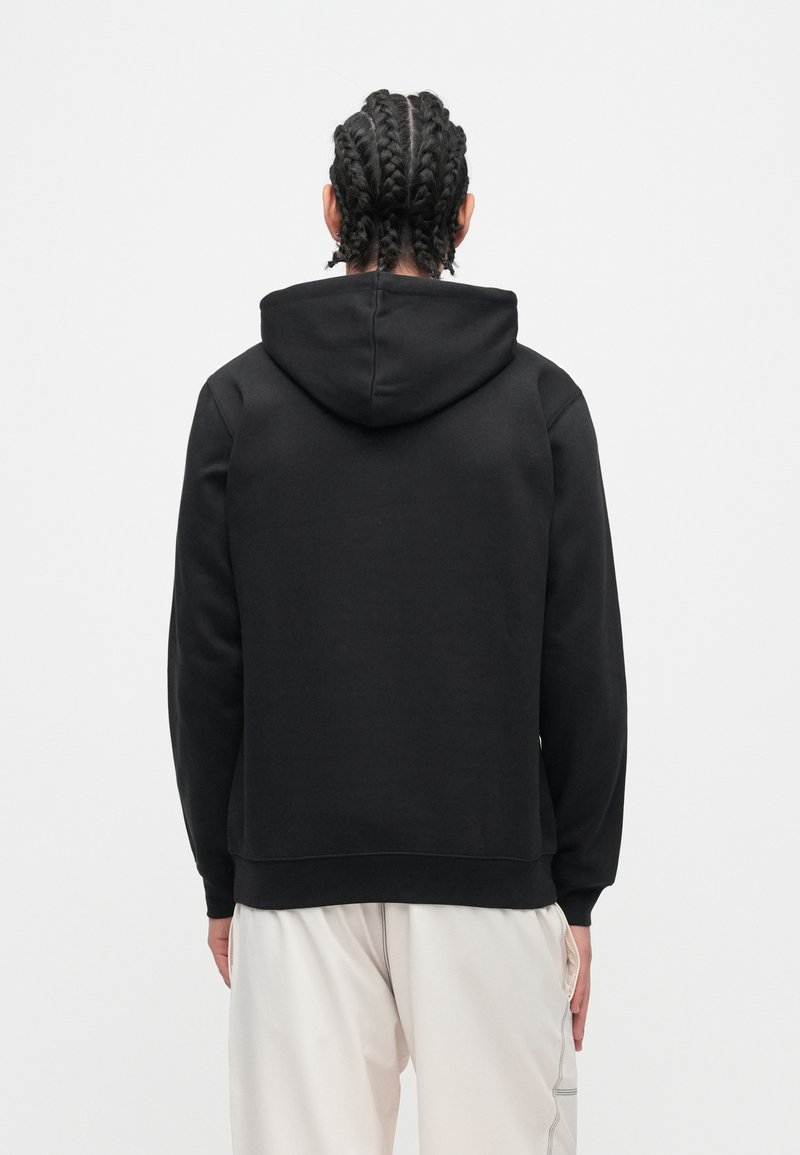 Persona con capelli intrecciati che indossa una felpa con cappuccio nera e pantaloni chiari, di spalle su uno sfondo bianco semplice.