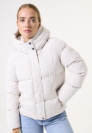 Doudoune beige clair avec un col haut, fermeture à boutons à l'avant et couture visible. Texture douce, deux poches latérales et détail de logo cousu.