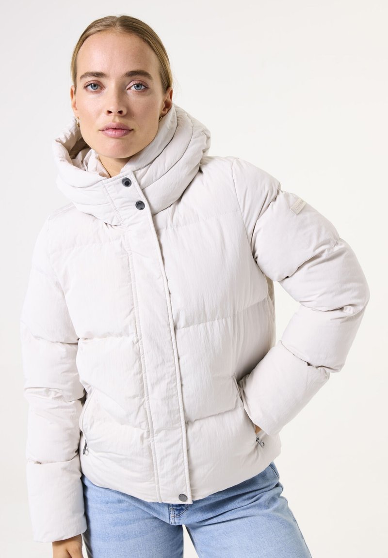 Chaqueta acolchada beige claro con cuello alto, cierre de botones en el frente y costuras visibles. Textura suave, dos bolsillos laterales y detalle de logo cosido.