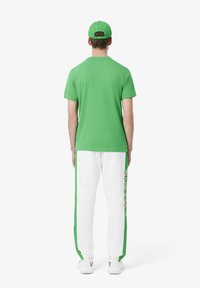 Grünes Baumwoll-T-Shirt, kombiniert mit weißen Hosen, die grüne Seitenstreifen und das "LACOSTE"-Logo aufweisen. Die Accessoires beinhalten eine passende grüne Kappe.