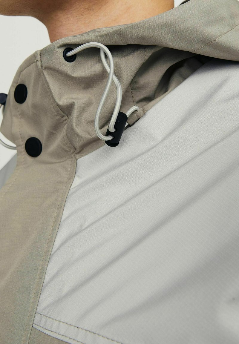 Lichte jack met een beige en grijze kleurenschema, met een textuurafwerking, twee zwarte knopen en verstelbare trekkoordhardware bij de kraag.