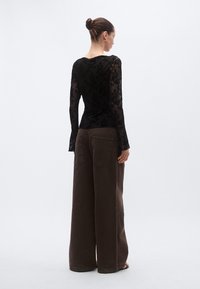 Črna čipkasta majica z dolgimi rokavi in razširjenimi manšetami, združena z rjavimi širokimi hlačami z zadnjimi žepi. Teksturiran, večplastni dizajn.
