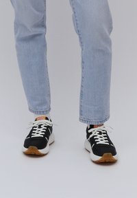 Schwarze Sportschuhe mit weißen Schnürsenkeln, ausgestattet mit einer weißen Zwischensohle und einer braunen Gummisohle. Getragen mit hellblauen Jeans und weißen Socken.