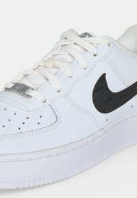 Άσπρο αθλητικό παπούτσι Nike με μαύρο σήμα, λευκά κορδόνια, διάτρητη μύτη και μεταλλική ετικέτα κορδονιών που δείχνει "AF1" κοντινή εικόνα.