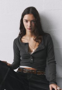 Grau gestreiftes Langarm-Oberteil mit Knopfleiste und tiefem Ausschnitt, über einem hellen Unterhemd getragen. Kombiniert mit dunkelblauen Jeans und einem mit Nieten besetzten Gürtel.