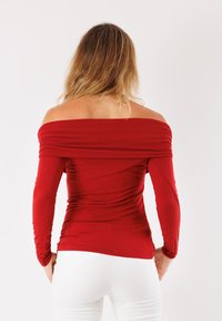 Top rosso scollato con spalle scoperte, maniche lunghe, design aderente e texture arricciata sul collo e in vita. Tessuto elastico e liscio.