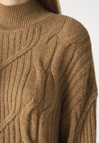 Ruskea neulepusero, jossa on korkea kaulus, ribattu ja kuvioitu pinta, jonka viimeistely on tekstuuriutunut ja pehmeä ulkonäkö.