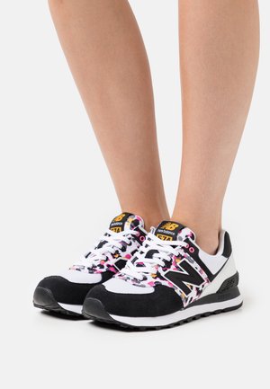 Jalat, joissa on valkoiset ja mustat New Balancen lenkkarit, joissa on vaaleanpunaiset ja oranssit leopardikuvioiset yksityiskohdat, valkoiset nauhat ja keltainen logo kielen päällä.