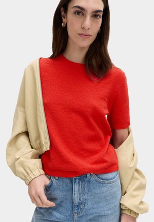 Vrouw met donker haar draagt een rood kort mouwtruitje, licht beige jas hangend over de schouders en een blauwe spijkerbroek tegen een egale achtergrond.