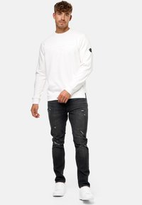 Witte sweatshirt met reliëflogo, gecombineerd met versleten zwarte jeans en witte sneakers. De stof lijkt zacht met een ontspannen pasvorm.