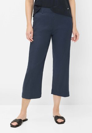 Persoon draagt marineblauwe wijde cropped broek, zwarte instapsandalen en een zwart topje, staand tegen een effen witte achtergrond.