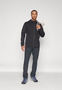 Svart zip-up jakke med quiltet front, hette og to sidelommer, kombinert med mørke bukser og grå-blå sko, modell som holder en beige gjenstand.