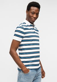 Νέος άνδρας που φοράει λευκό πόλο με μπλε και μαύρες οριζόντιες ρίγες, στέκεται με τα χέρια στις τσέπες του ανοιχτό μπλε τζιν.