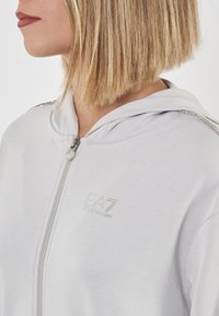 Felpa con zip grigia chiara presenta una consistenza morbida, un cappuccio e dettagli argentati sulle spalle. Logo EA7 mostrato sul davanti.