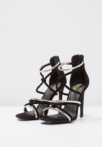 Sandales à talons hauts en daim noir avec accents dorés, présentant plusieurs fines lanières et un design noué. Bout ouvert et fermeture éclair au dos.