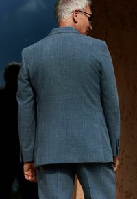 Chaqueta de traje de textura azul claro con un ajuste ceñido, que presenta un cuello con muescas, dos botones y detalles de costura sutiles.