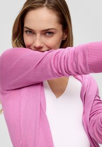 Różowy, dziergany kardigan z prążkowaną teksturą i długimi rękawami, noszony na białym, prążkowanym topie z dekoltem w serek. Zbliżenie na ramię.
