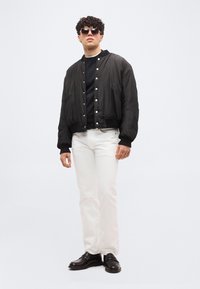 Νεαρός άνδρας που φοράει μαύρο μπουφάν bomber, μαύρο πουλόβερ, λευκό παντελόνι, μαύρα μοκασίνια και γυαλιά ηλίου, στέκεται μπροστά από έναν απλό φωτεινό φόντο.