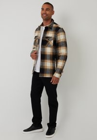 Kariertes Flanellhemd in Creme, Schwarz und Braun mit zwei Brusttaschen, kombiniert mit schwarzen Hosen und schwarzen Sneakers, strukturierter Stoff.