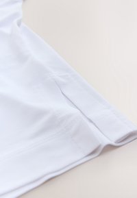 Gros plan sur l’ourlet d’un vêtement en tissu blanc avec une petite fente et des coutures visibles sur un fond clair.