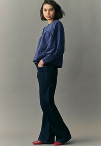 Blouse en coton bleu marine à manches longues avec des épaules froncées, associée à un jean flare sombre et des ballerines rouges. Le mannequin se tient les mains dans les poches.