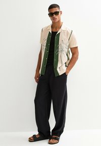 Camicia a maniche corte con motivo a palme in beige e verde, abbinata a pantaloni neri a gamba larga e sandali neri. Gli accessori includono occhiali da sole scuri.