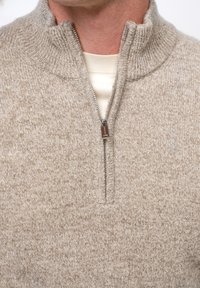 Beżowy sweter, teksturowany materiał dzianinowy, pół zamek przy szyi z srebrnymi akcentami, noszony na kremowej koszuli.