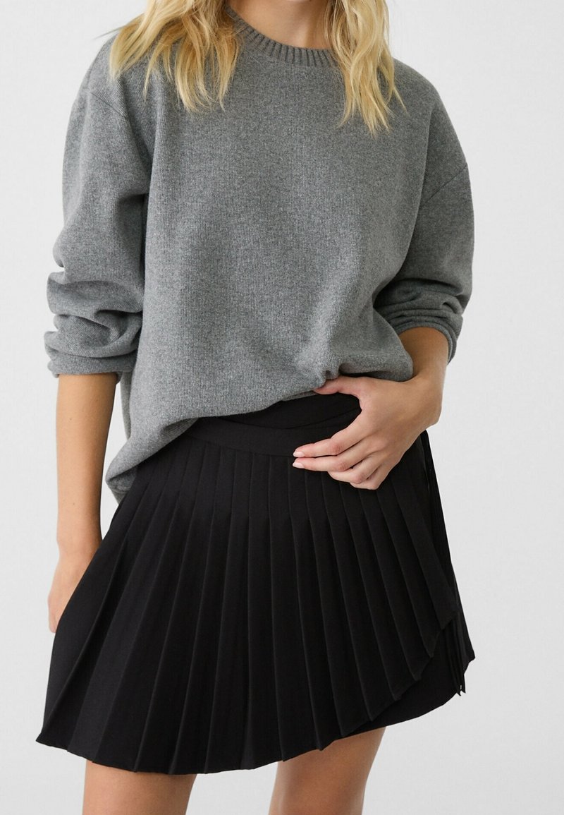 Pull en tricot gris à manches longues, associé à une jupe noire plissée avec une taille haute et un design fluide.