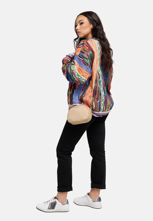 Vrouw met een kleurrijke gebreide trui met patroon, zwart opgerolde broek, witte sneakers en een beige schoudertas, poseert zijwaarts tegen een witte achtergrond.