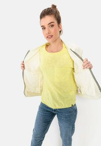 Gelbes, leichtes T-Shirt mit einer Vorderstasche, kombiniert mit blauen Jeans und einer offenen weißen Jacke, die ein fusseliges Innenfutter zeigt.