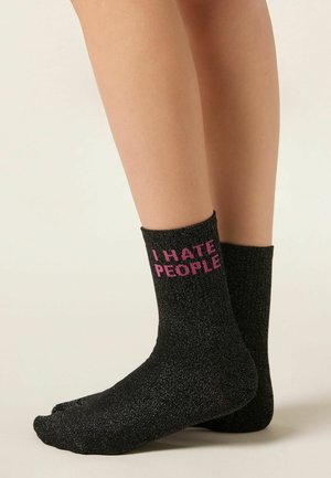 Feet wearing black glittery socks with pink text reading "I HATE PEOPLE," on a plain light background.