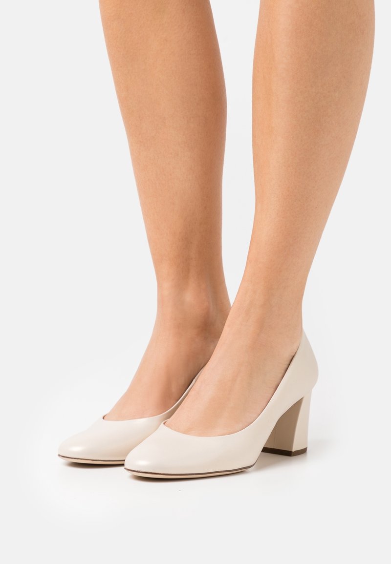 Pieds portant des chaussures à petit talon carré de couleur blanc cassé sur fond blanc, montrant une peau lisse et des chevilles.