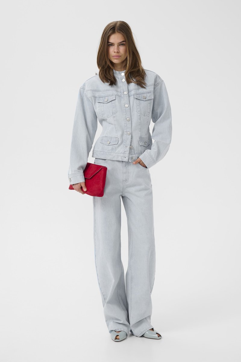 Nő világoskék farmerdzsekiben és bő szárú farmerral, piros borítéktáskát tartva, egyszínű fehér háttér előtt áll.