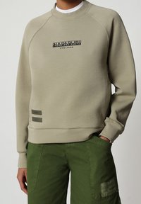 Sudadera beige con cuello redondo, mangas raglán y un estampado de logo negro. Presenta etiquetas de acento grises en el dobladillo. Combinada con pantalones verdes.