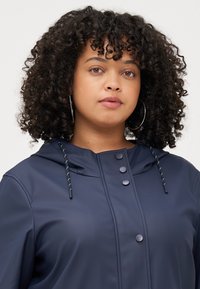 A young woman with curly hair wears a navy hooded jacket, gazing directly at the camera with a neutral expression.
