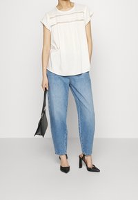 Donna che indossa una blusa bianca con dettagli in pizzo, jeans azzurri chiari e scarpe con tacco nere, con una borsa nera al suo fianco.