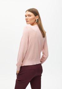 Rosa långärmad tröja med rundad halsringning och ribbad nederkant. Modellen bär vinröda byxor; bakgrunden är helt vit.