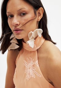 Pfirsichfarbiges Halterkleid mit floraler Spitze am Ausschnitt, aus transparentem Stoff und auffälligen Statement-Ohrringen mit schichtigen, bandartigen Details.