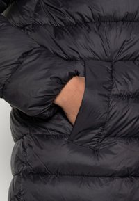 Zwarte pufferjack met gewatteerd ontwerp, zachte glanzende textuur en zijzak. De stof lijkt lichtgewicht en geïsoleerd.