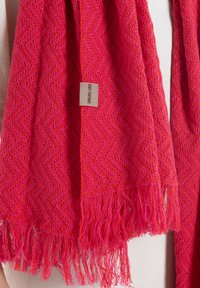 Roze gebreide sjaal met een visgraatpatroon, voorzien van franjes aan de randen en een klein label met tekst.