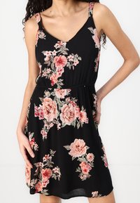 Robe noire sans manches avec des motifs floraux roses et pêche, de longueur genou, portée par une femme avec la main sur la hanche.