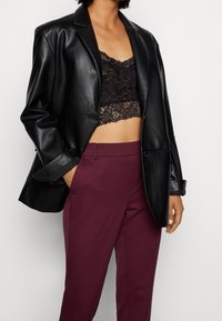 Czarna sztuczna skórzana marynarka o dopasowanym kroju, noszona na czystym koronkowym biustonoszu w kolorze czarnym, w zestawieniu z burgundowymi spodniami o prostym kroju.