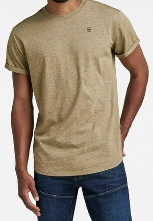 Homme portant un t-shirt à manches courtes marron clair avec les manches retroussées et un jean bleu foncé, sur fond blanc.