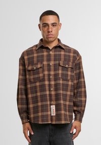 Brown plaid flannel shirt with two chest pockets, button-down front, and a woven label at the hem. Soft texture, relaxed fit, long sleeves.