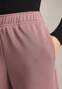 Világos rózsaszín tréningnadrág puha anyagból, elasztikus derékpánttal és oldalzsebekkel, semleges háttér előtt bemutatva.