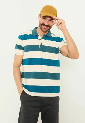 Mann lächelt, trägt senffarbene Mütze, türkis-weiß gestreiftes Poloshirt, schwarze Hose, mit der Hand die Mützenkrempe vor schlichtem Hintergrund berührend.